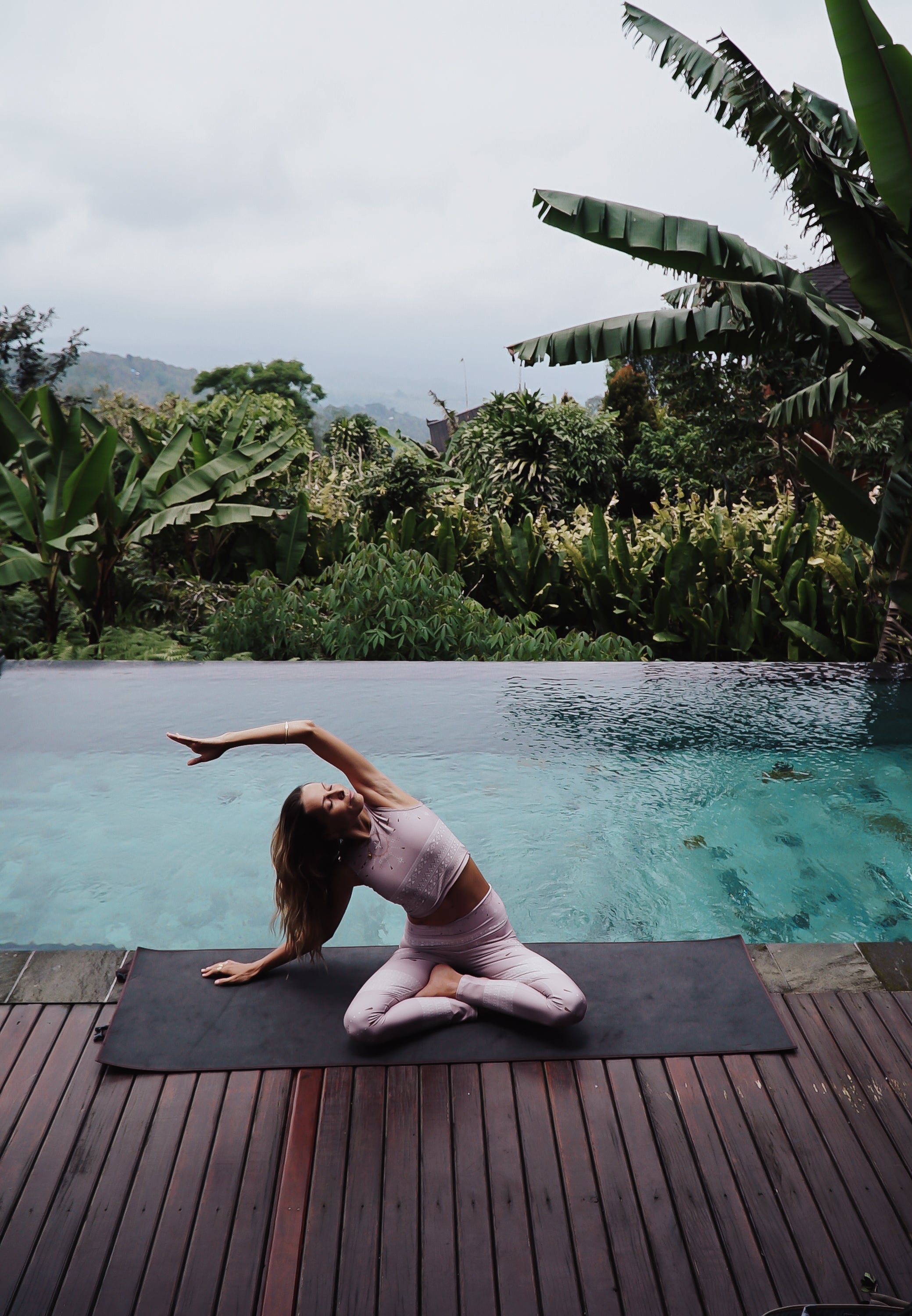 woman doing yoga on the edge of an infintiy pool in the jungle - on a black yoga mat in a dusty pink yoga set