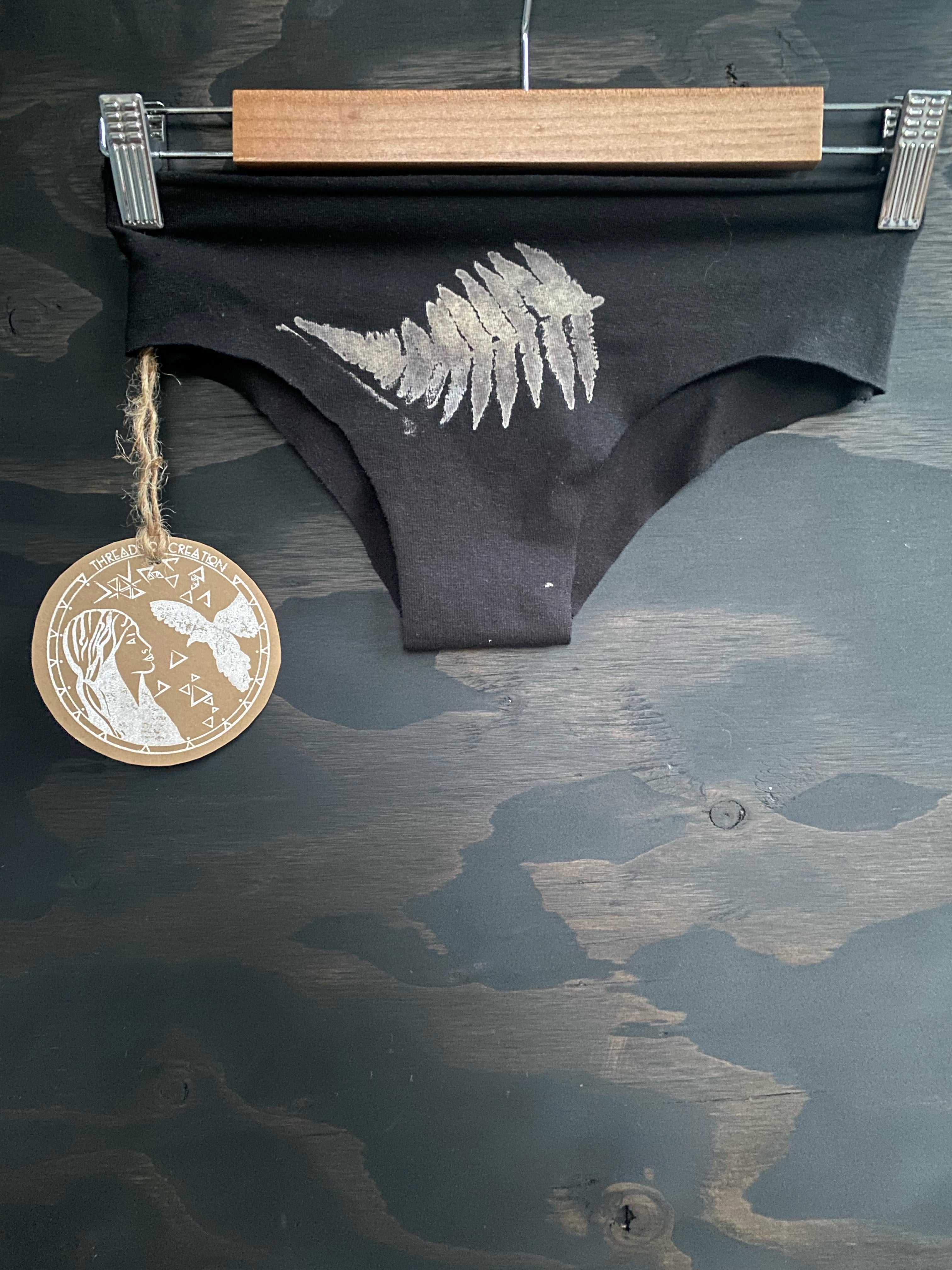 Black hemp panties with a printed fern design, displayed on a hanger, with a round sustainable cotton label below.