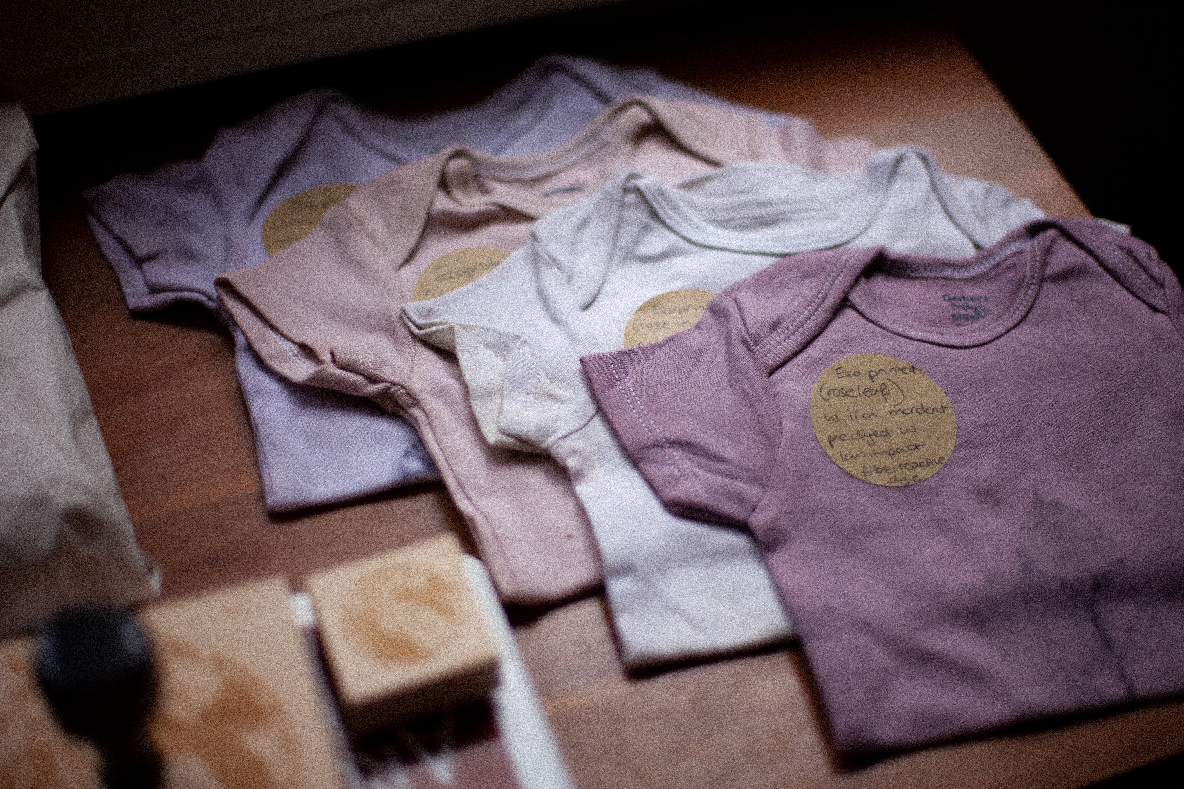 4 hand dyed and eco printed baby onesies in pastel shades folded on a wooden table