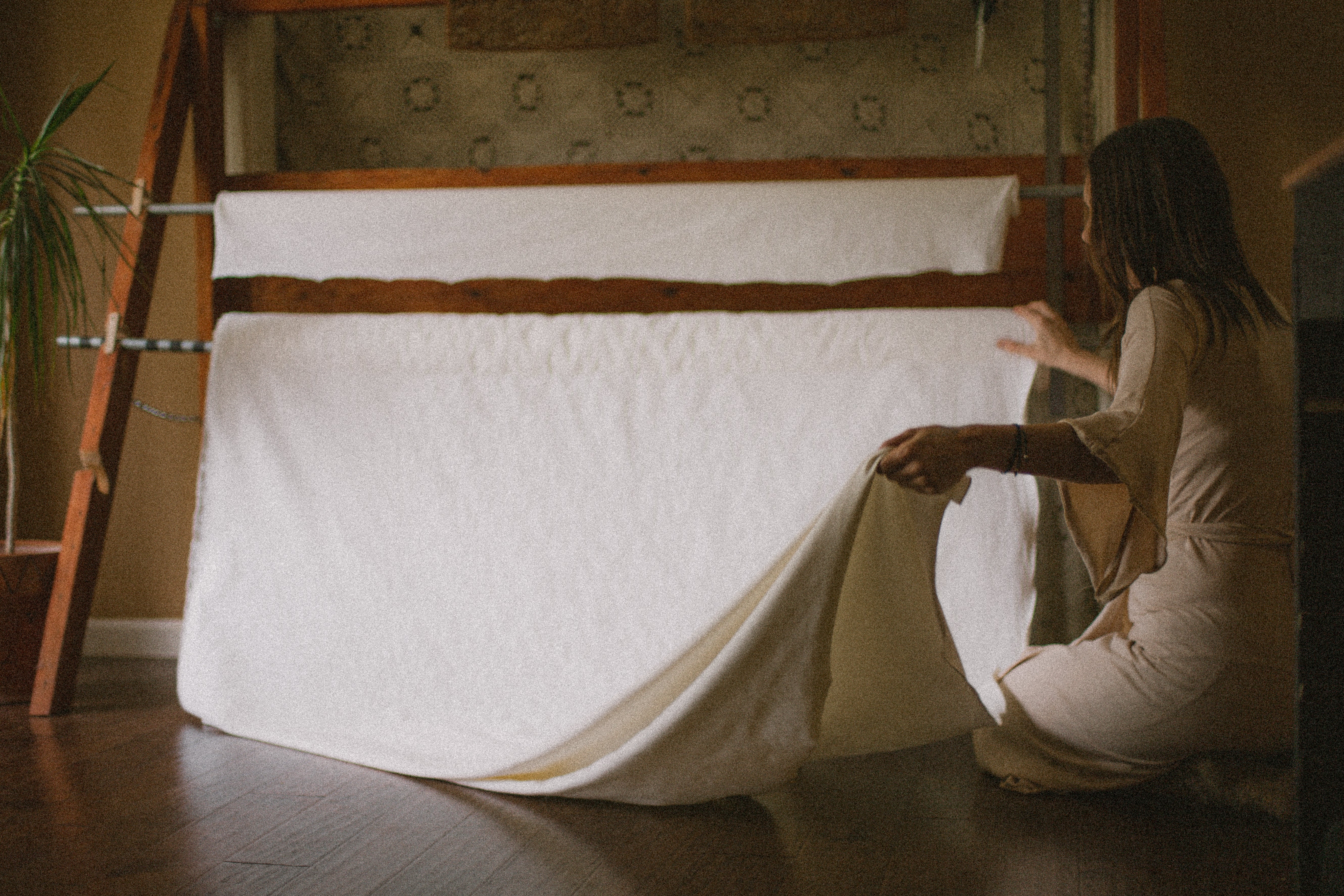 woman rolling out white fabric from a wooden fabric roll with a crochet background and a plant in the corner