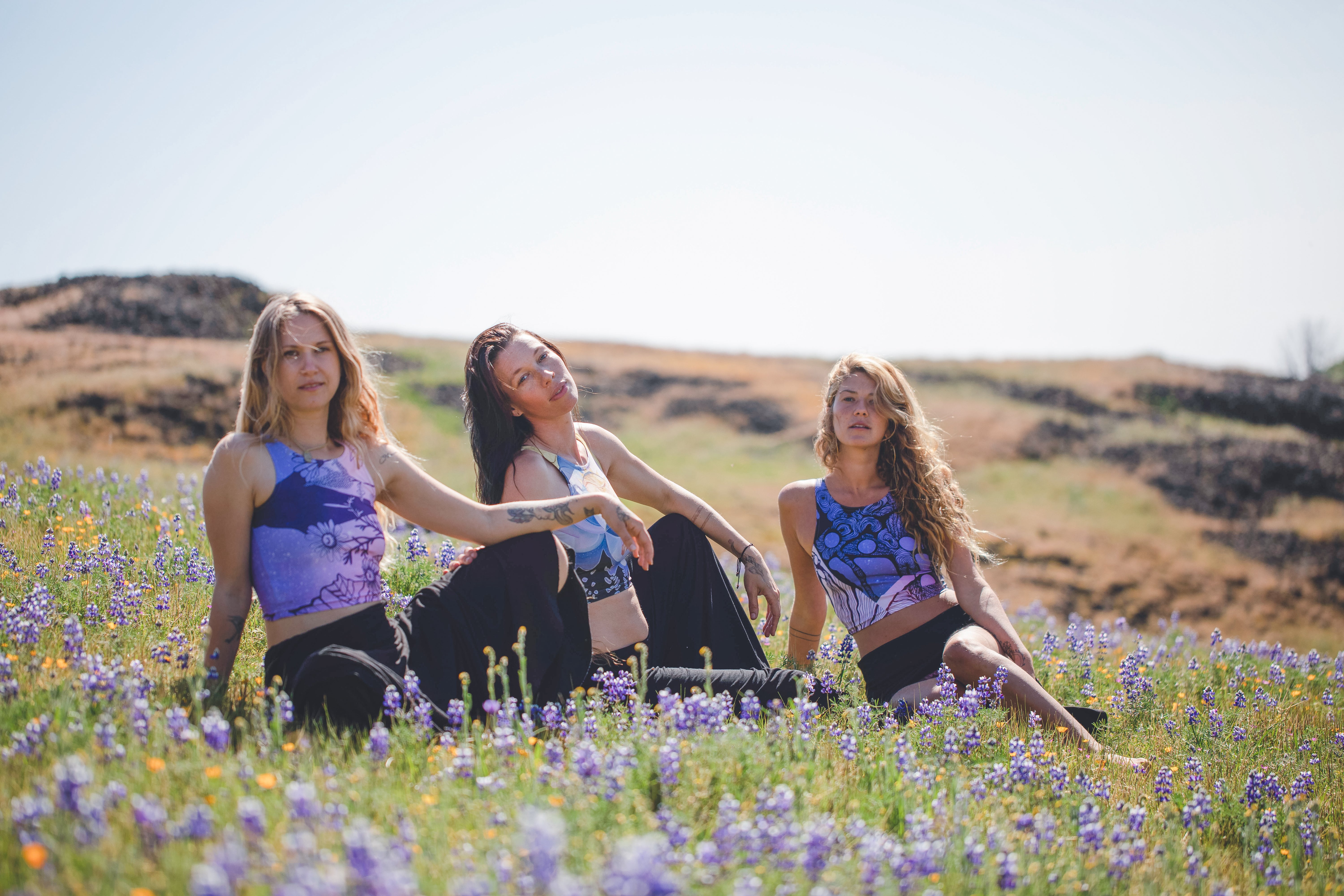 3 women wearing Yoga top in shades of pink and purple with plants in black in a field of flowers
