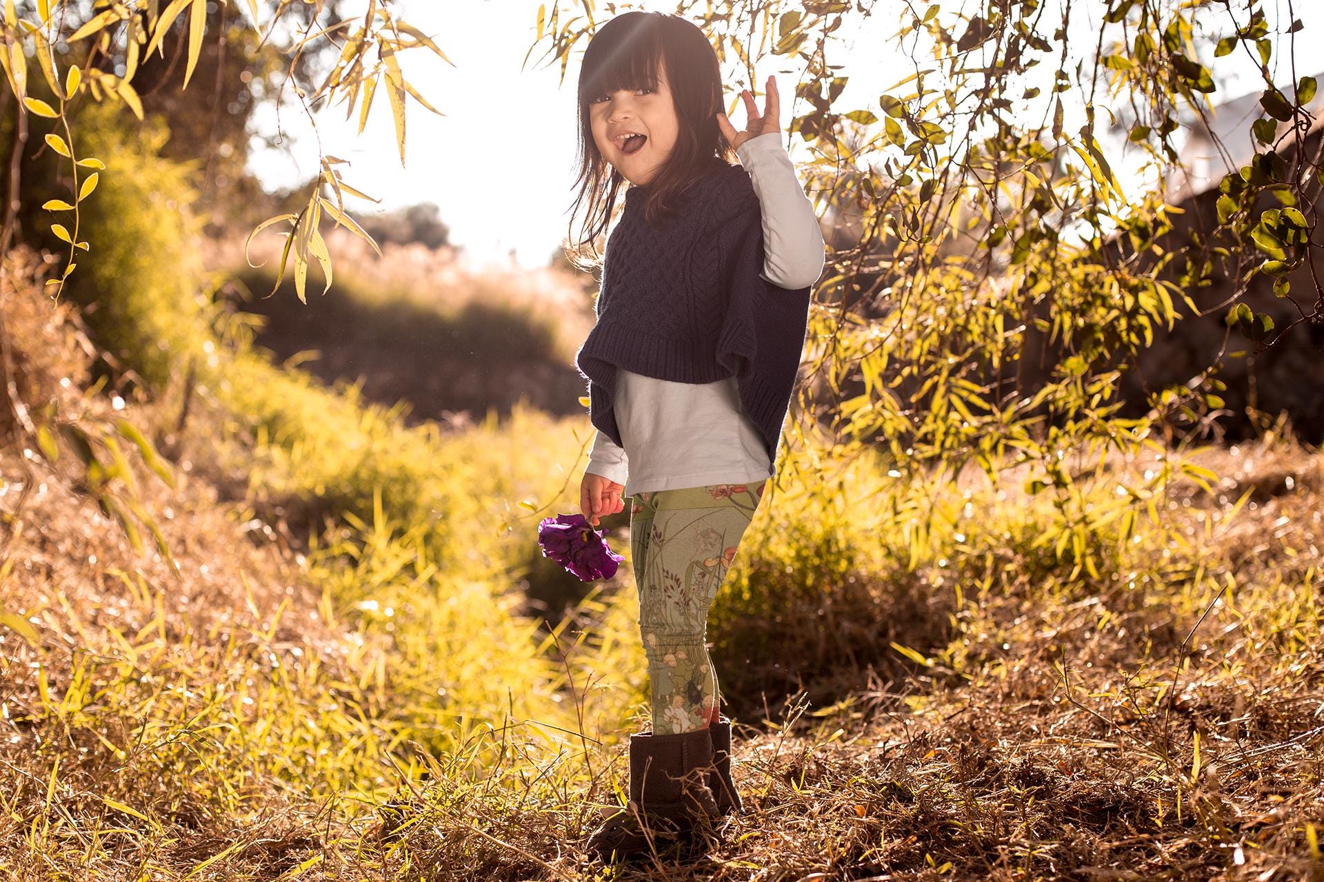 Little girl wearing green and blue illustrated leggings from Elsa beskows the root children. the leggings have images of little flower children in simple clothes and beautiful flowers. they are mostly teal green with some teal blue