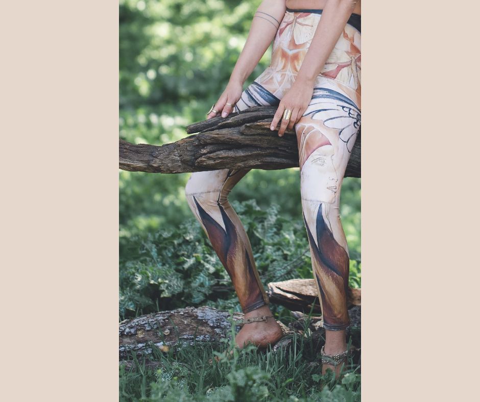 A person wearing multicolored leggings featuring artistic designs, including a silhouetted woman's face, raven wings, and butterfly patterns, seated on a tree stump in a natural outdoor setting.