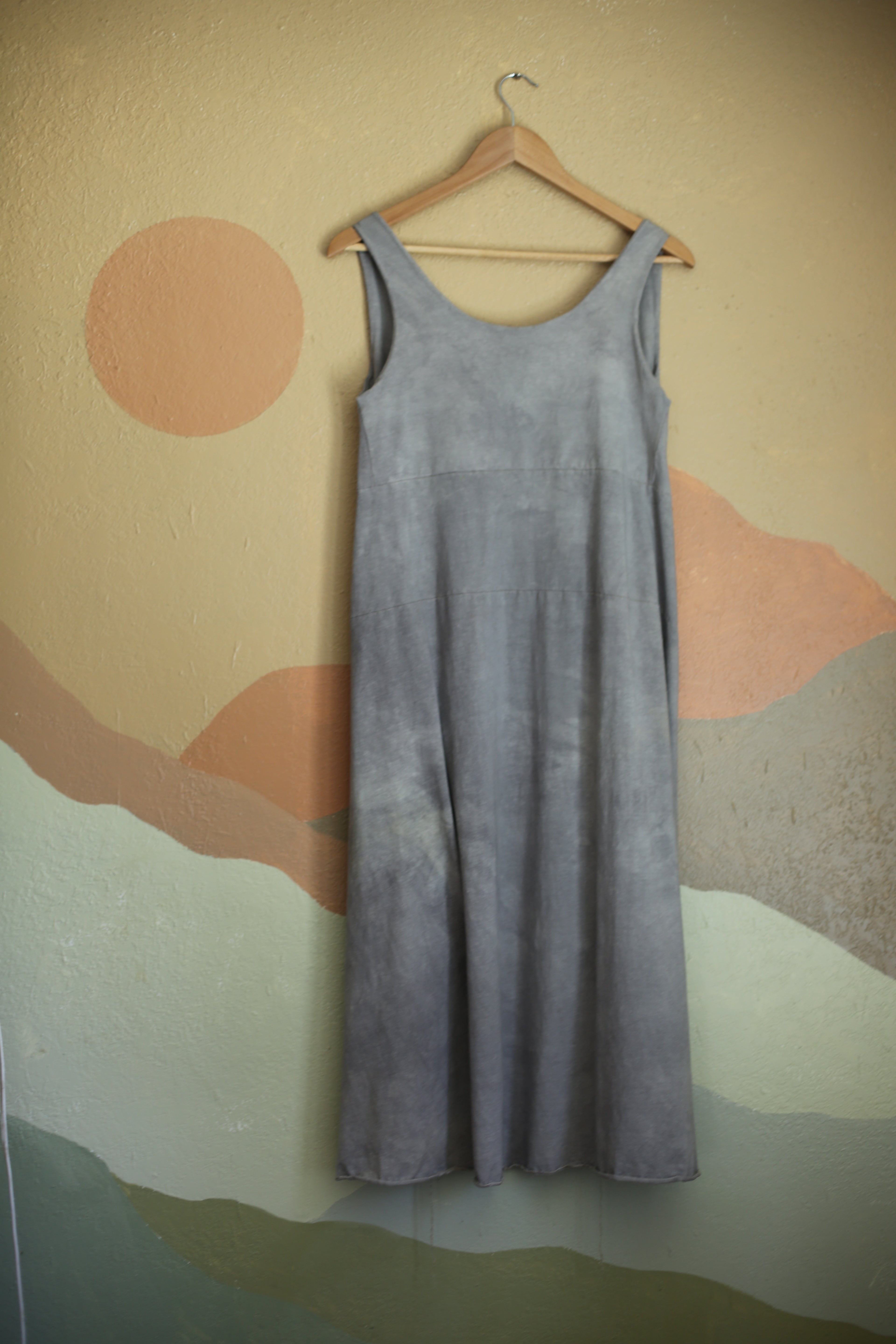 sleevless grey blue dress hanging on a wooden hanger against a back drop of a mural of the sun and mountains in pastel shades