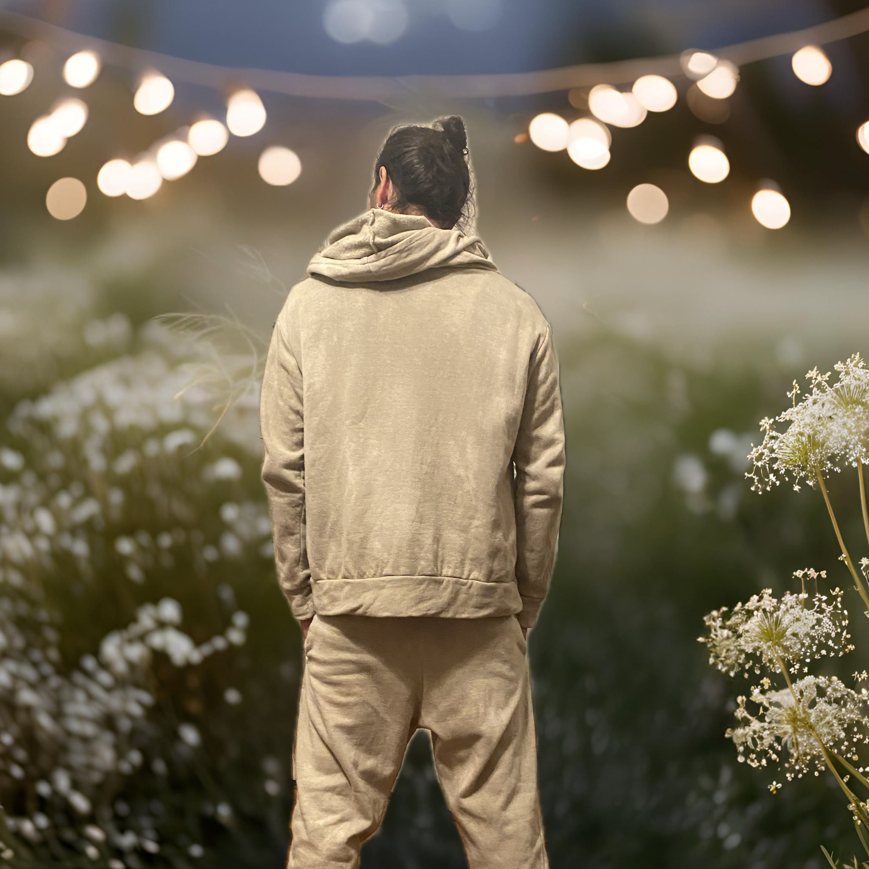 back view of a man with dark hair in a bun wearing a matching set of a hoody and sweat pants dyed in a shade of sage green with a ack drop of out of focus lights and flowers