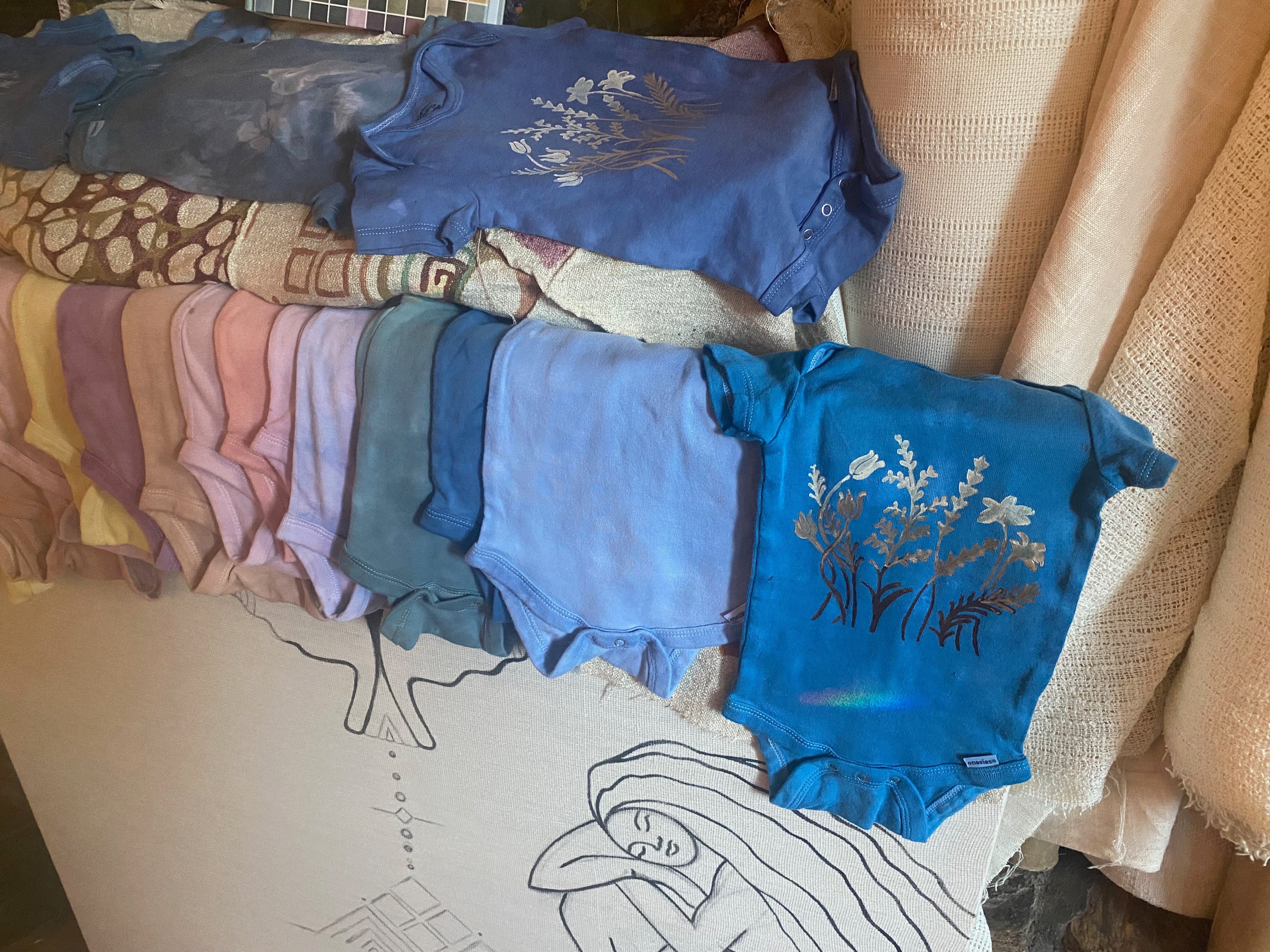 row of baby onesies in an array of bright hand dyed colors with flower screenprints on them 