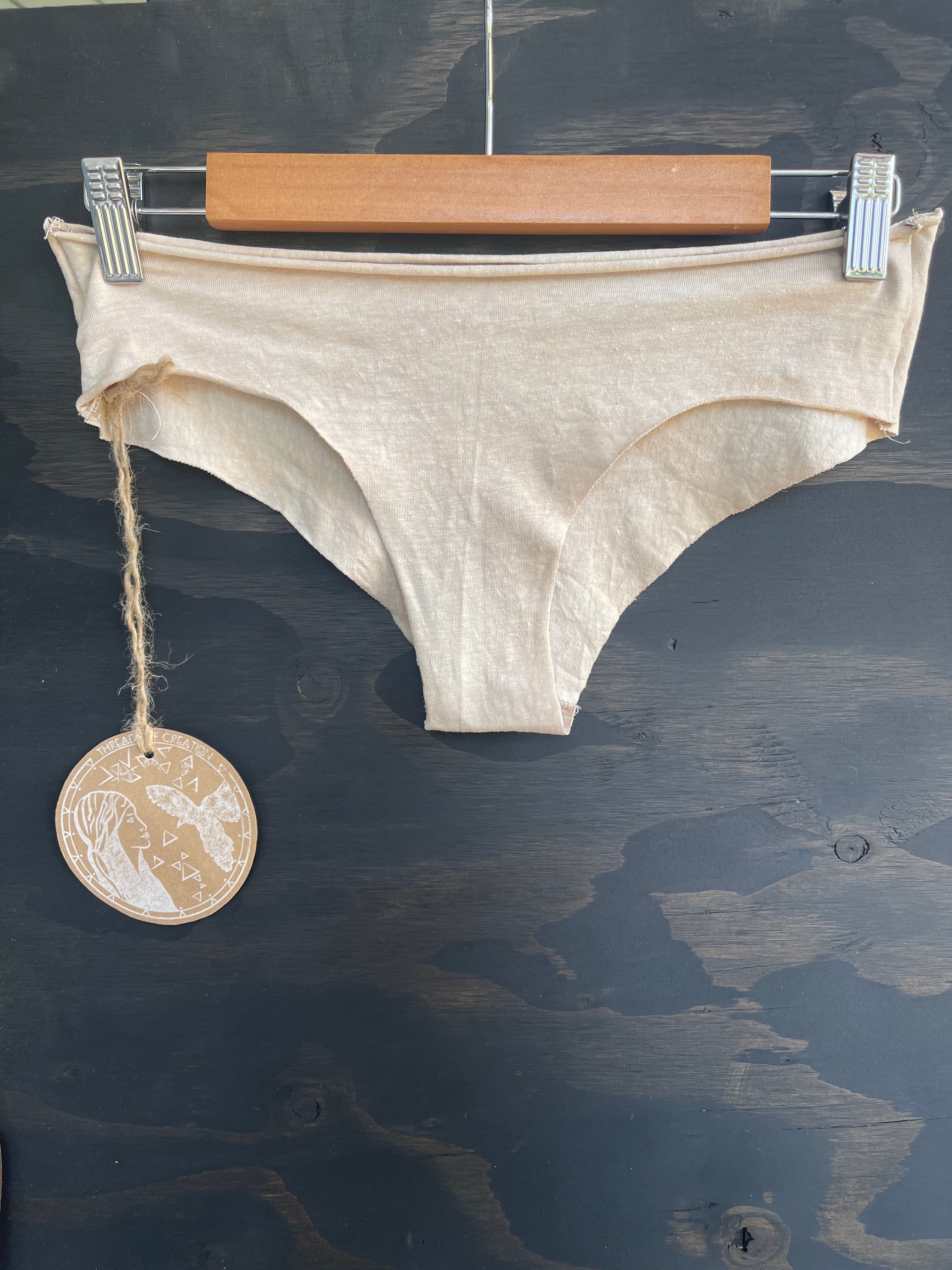 A pair of beige hemp panties displayed on a wooden hanger, with a round label showing a product code and the text 'Natural Dye'.