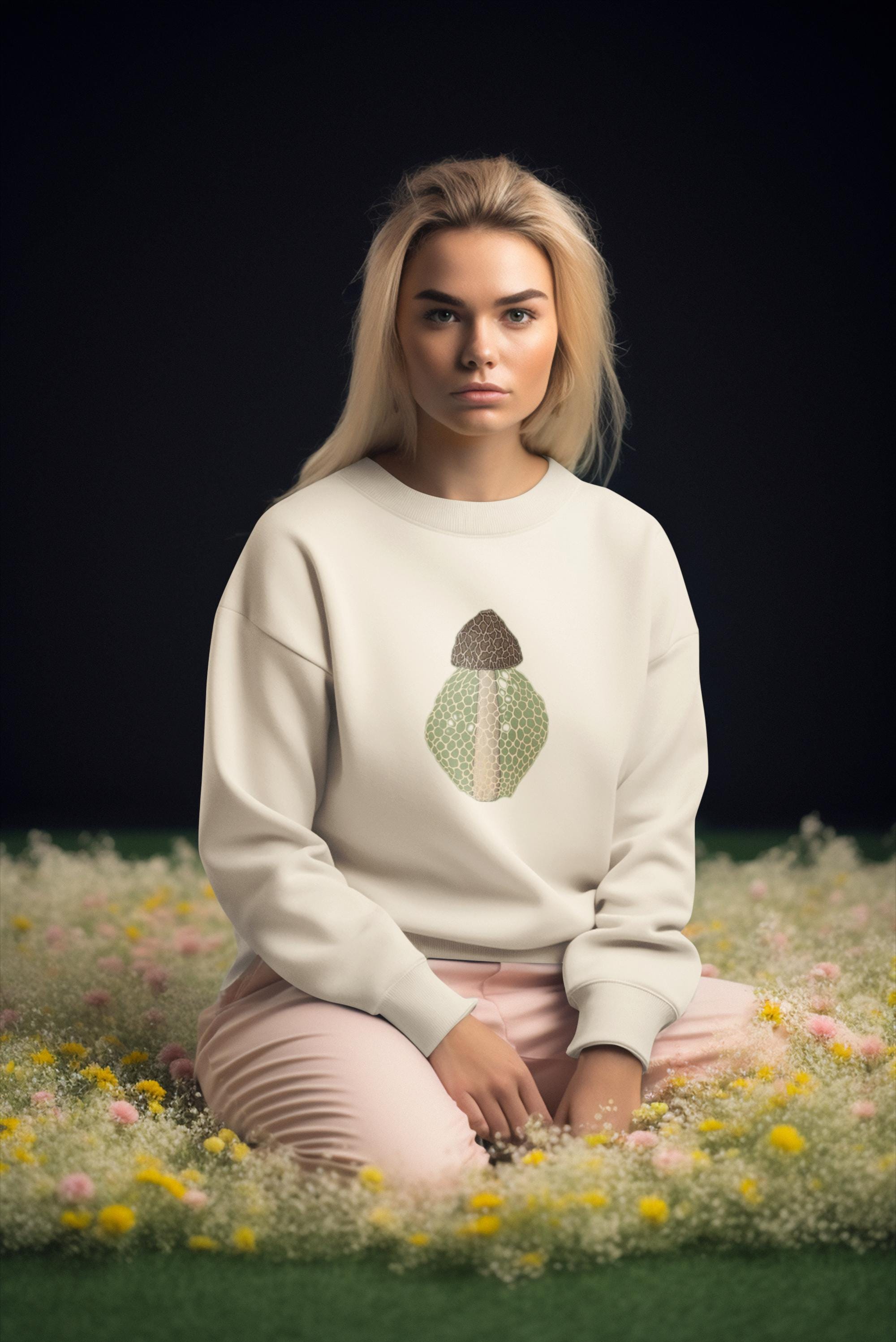 Beautiful woman with straight blonde hair sitting in a black room surrounded by pink flowers and green moss wearing a crew neck sweatshirt with a brown beige and green morel on it