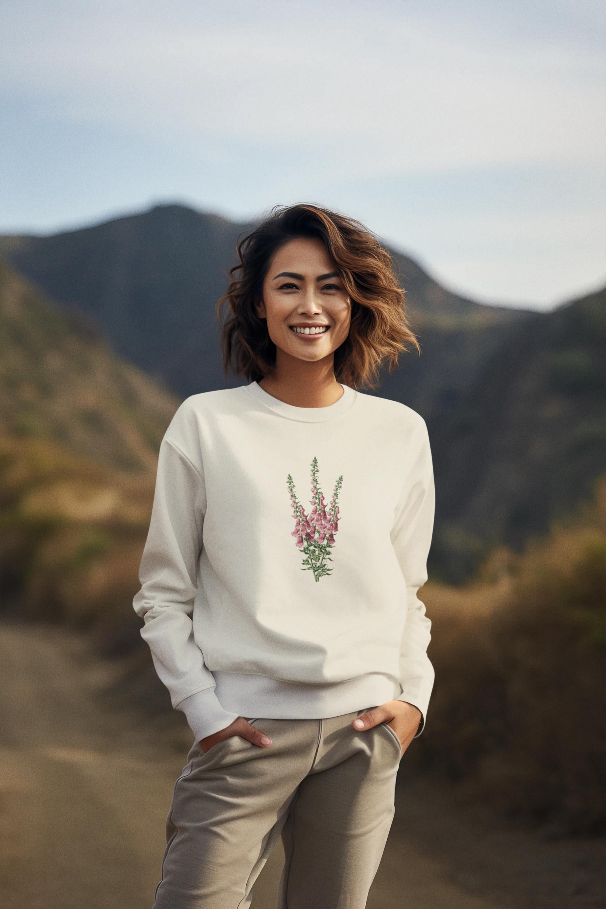 Young woman with short dark hair standing amidst a mountain range wearing a cream crewneck sweatshirt with a foxglove flower print on it