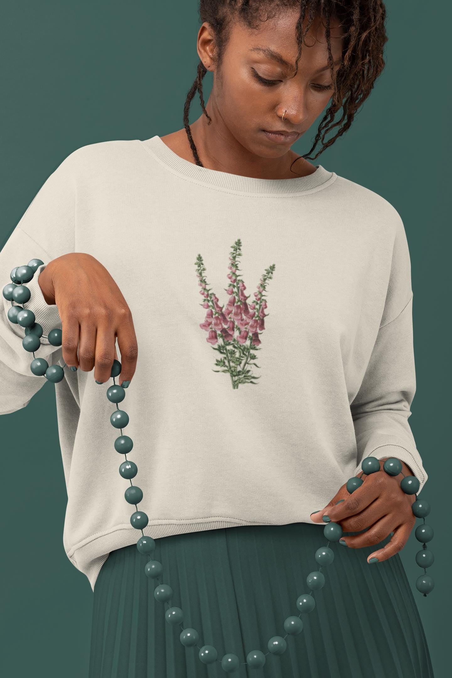 Young woman with dark brown hairl standing wearing a cream crewneck sweatshirt with a foxglove flower print on it and holding a green necklace