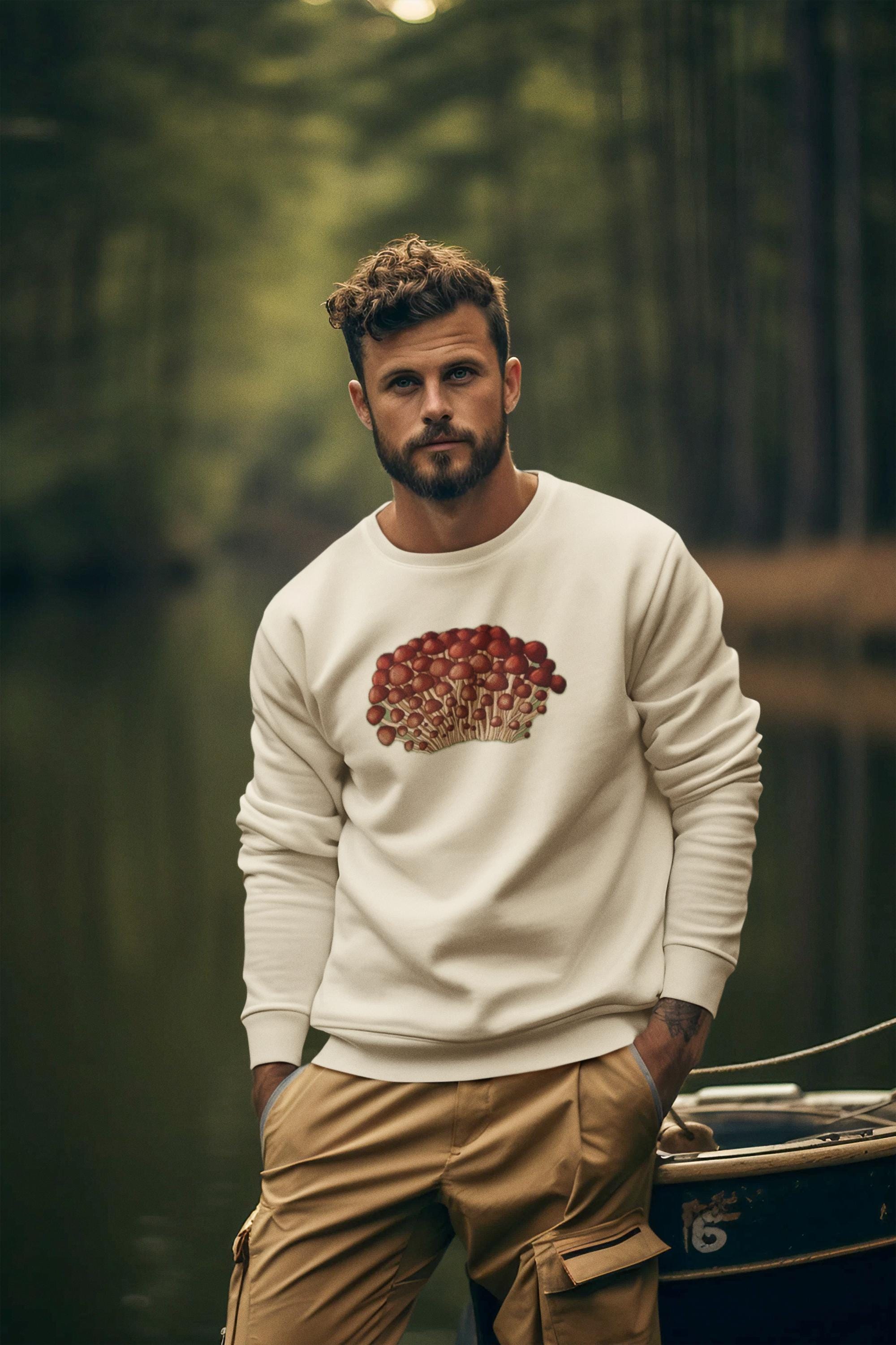 Beautiful man with brown wavey hair in the forest wearing a crewneck sweatshirt in cream with tiny little red and beige mushrooms