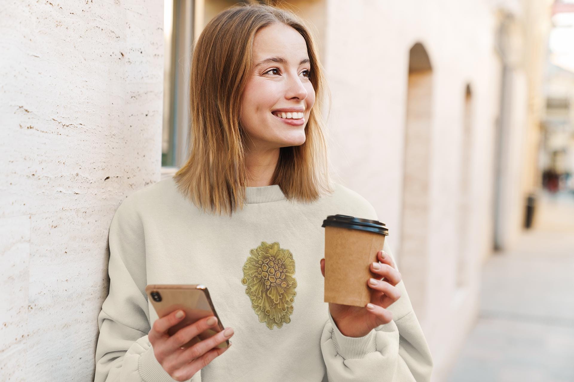 beautiful woman standing in the street holding a cup of coffee and looking at her phone. she has mid length blonde straight hair and she is wearing a cream crewneck sweater with yellow wavey mushrooms printed on it