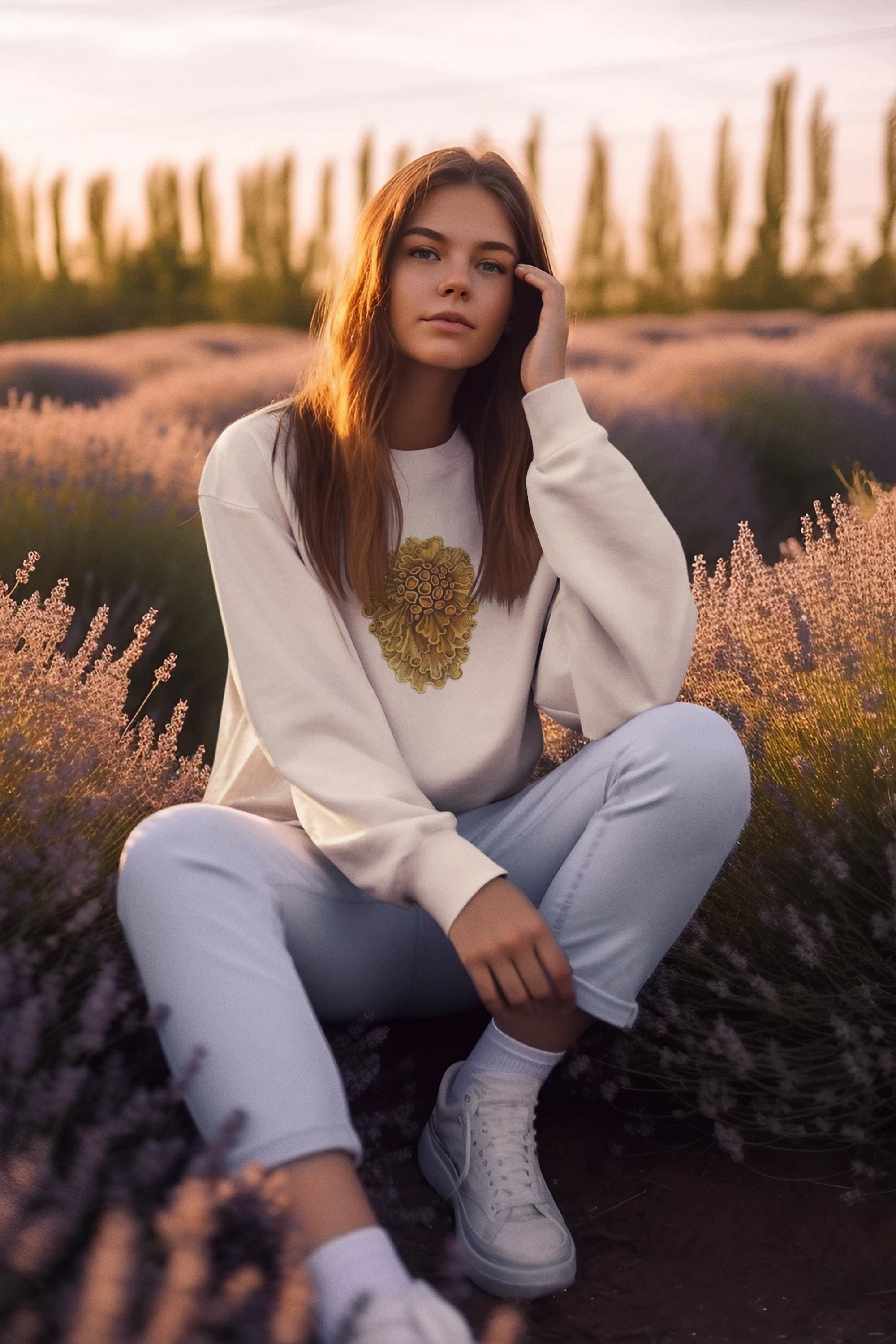 beautiful woman sitting  in the a lavender field. she has long brown wavey hair and she is wearing a cream crewneck sweater with yellow wavey mushrooms printed on it