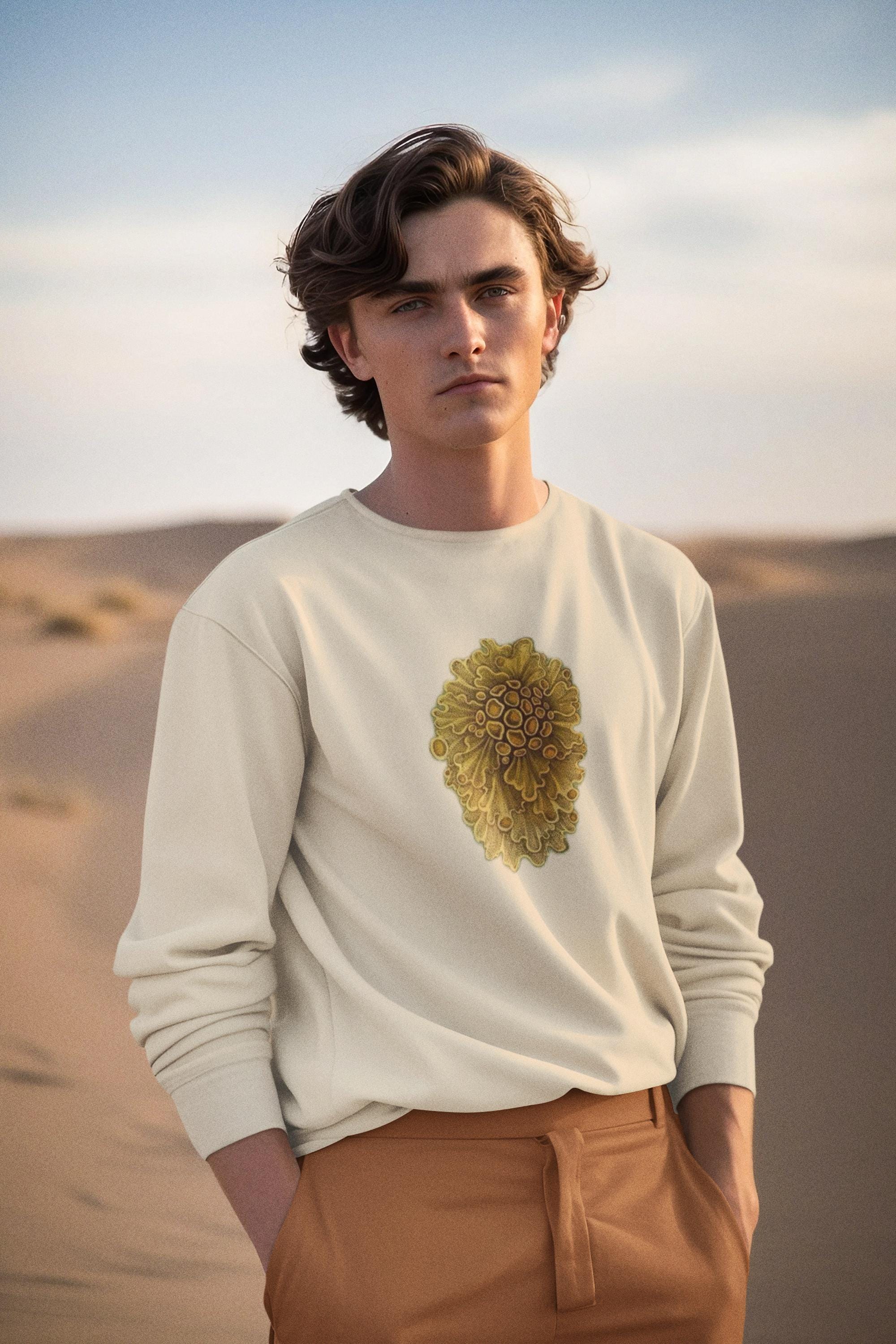 beautiful man standing in the desert. he has short brown wavey hair and she is wearing a cream crewneck sweater with yellow wavey mushrooms printed on it