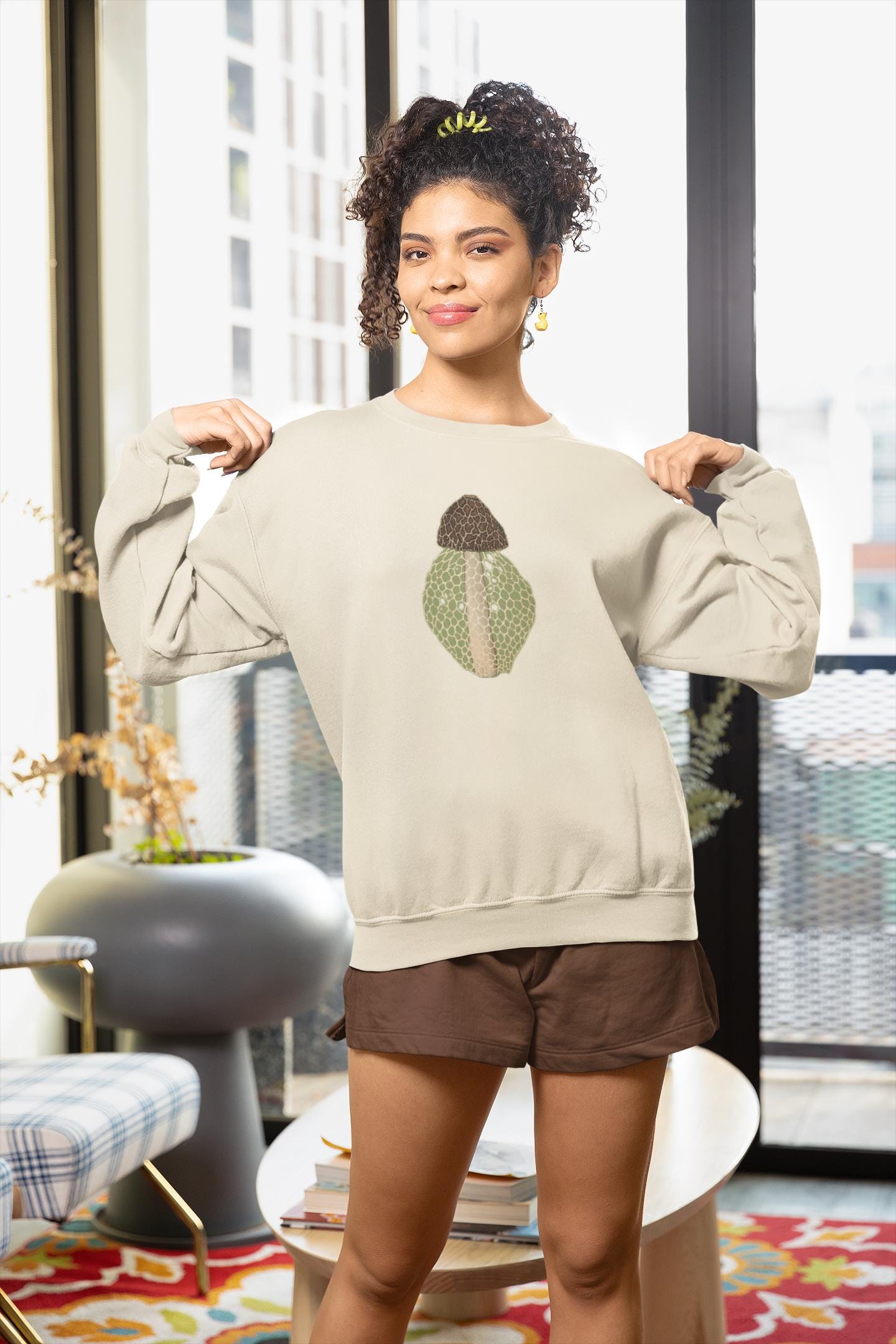 Beautiful woman with dark curly hair standing in an urban apartment building wearing a crew neck sweatshirt with a brown beige and green morel on it