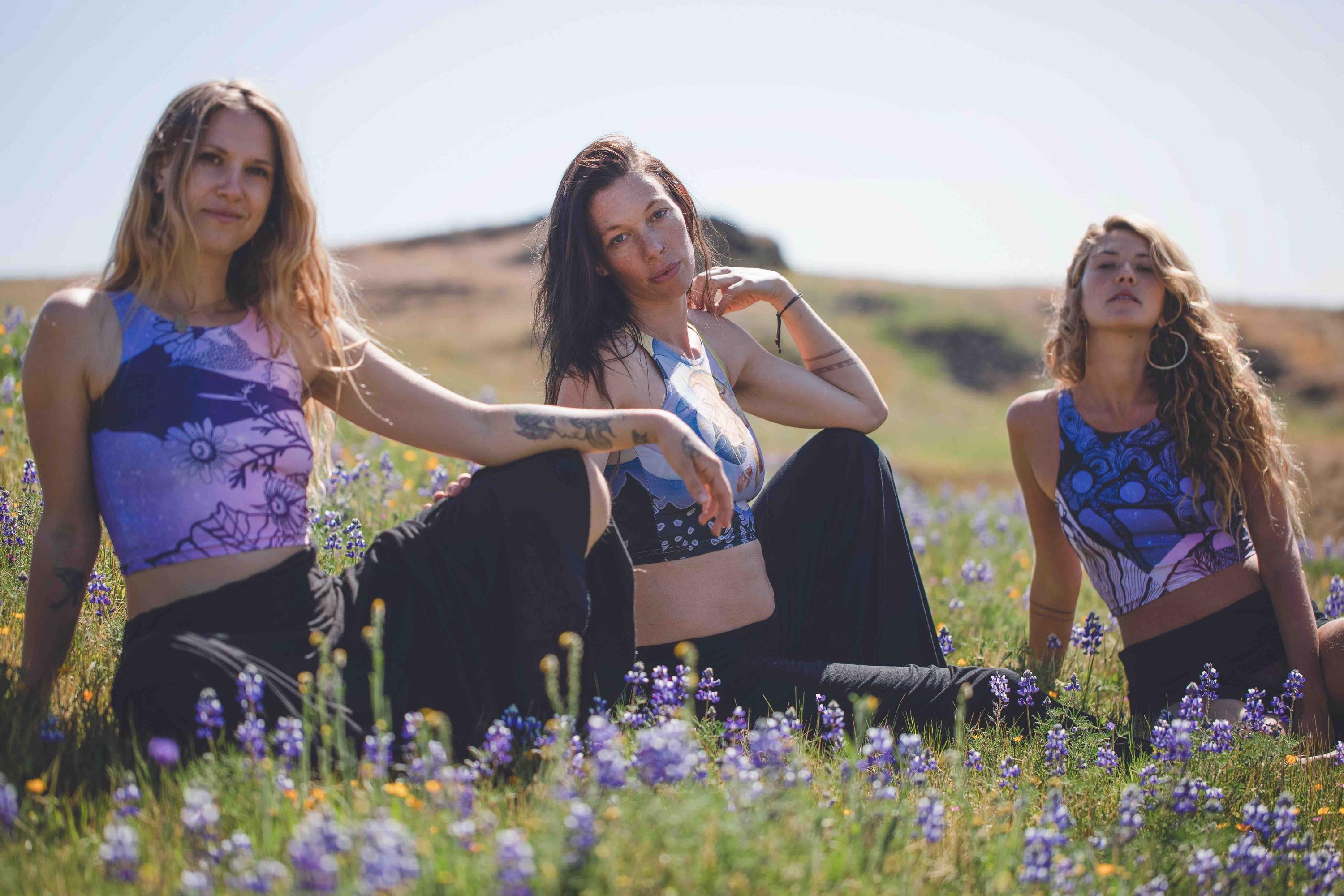 three women in field wearing Yoga tops in hues of purple and blue with a woman face in shades of gold and beige and geometric patterns