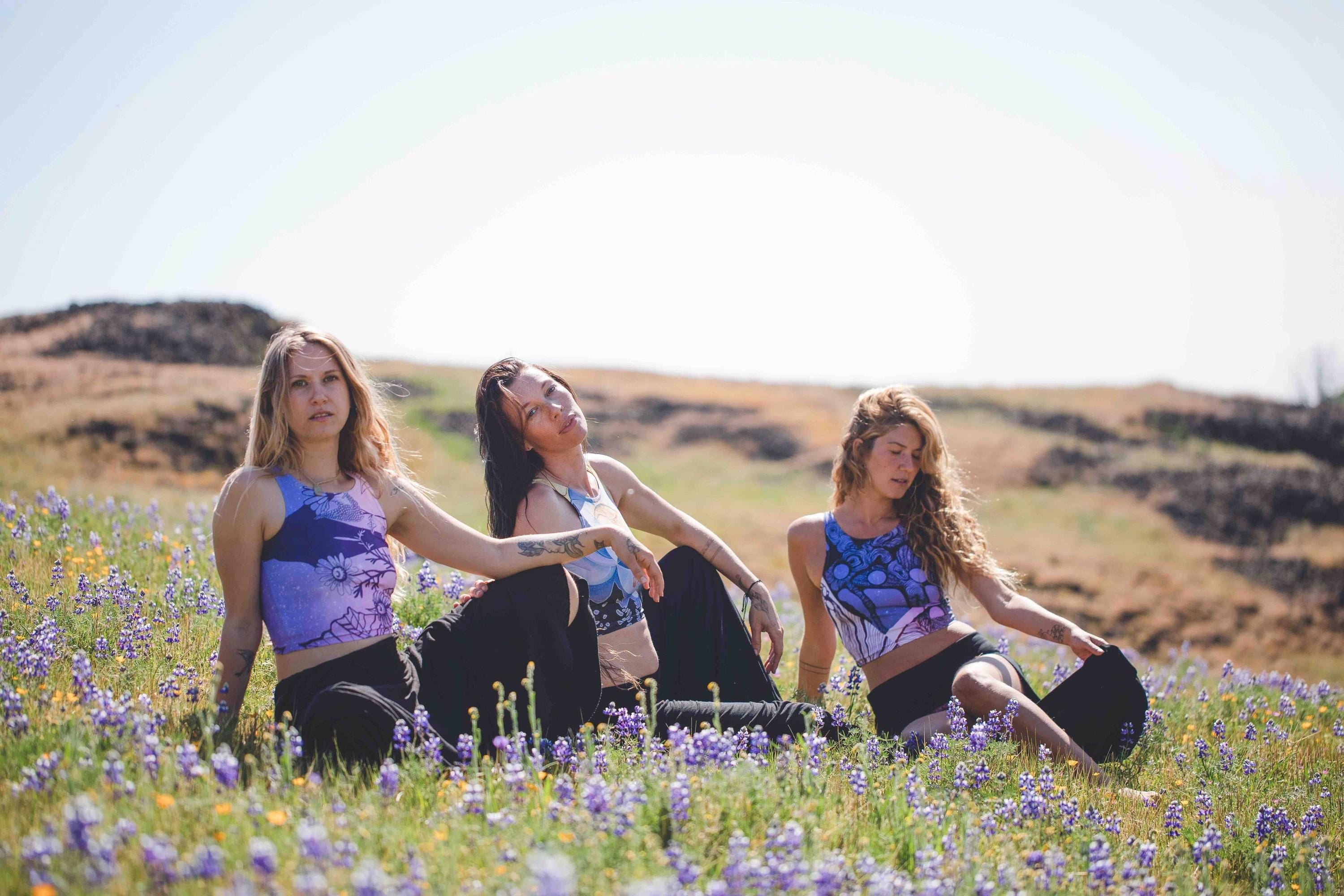 three women in field wearing Yoga tops in hues of purple and blue with a woman face in shades of gold and beige and geometric patterns