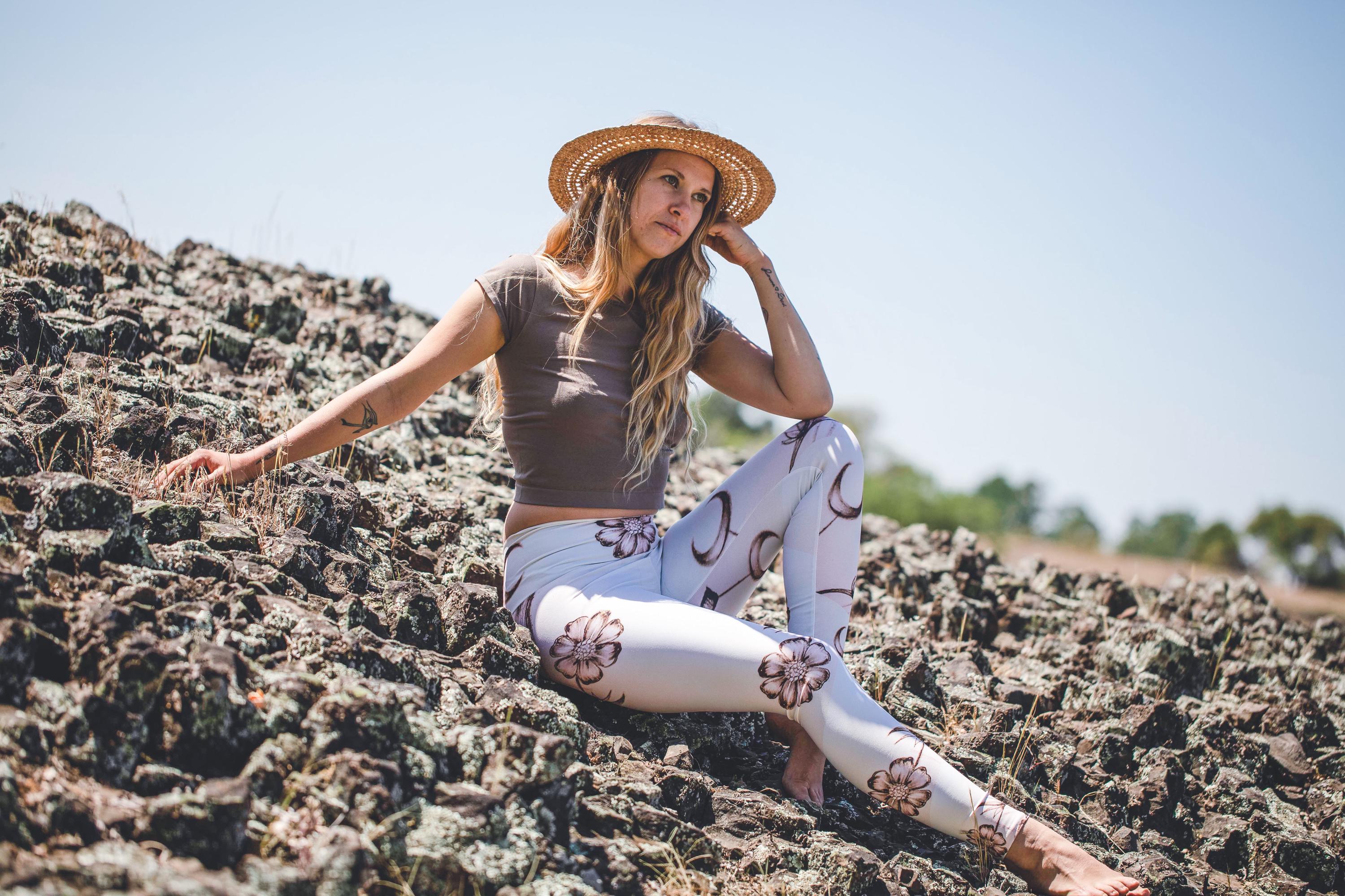 model sitting on hillside wearing Yoga leggings in shades of white and cream with images of flowers and crescent moons drawn on them