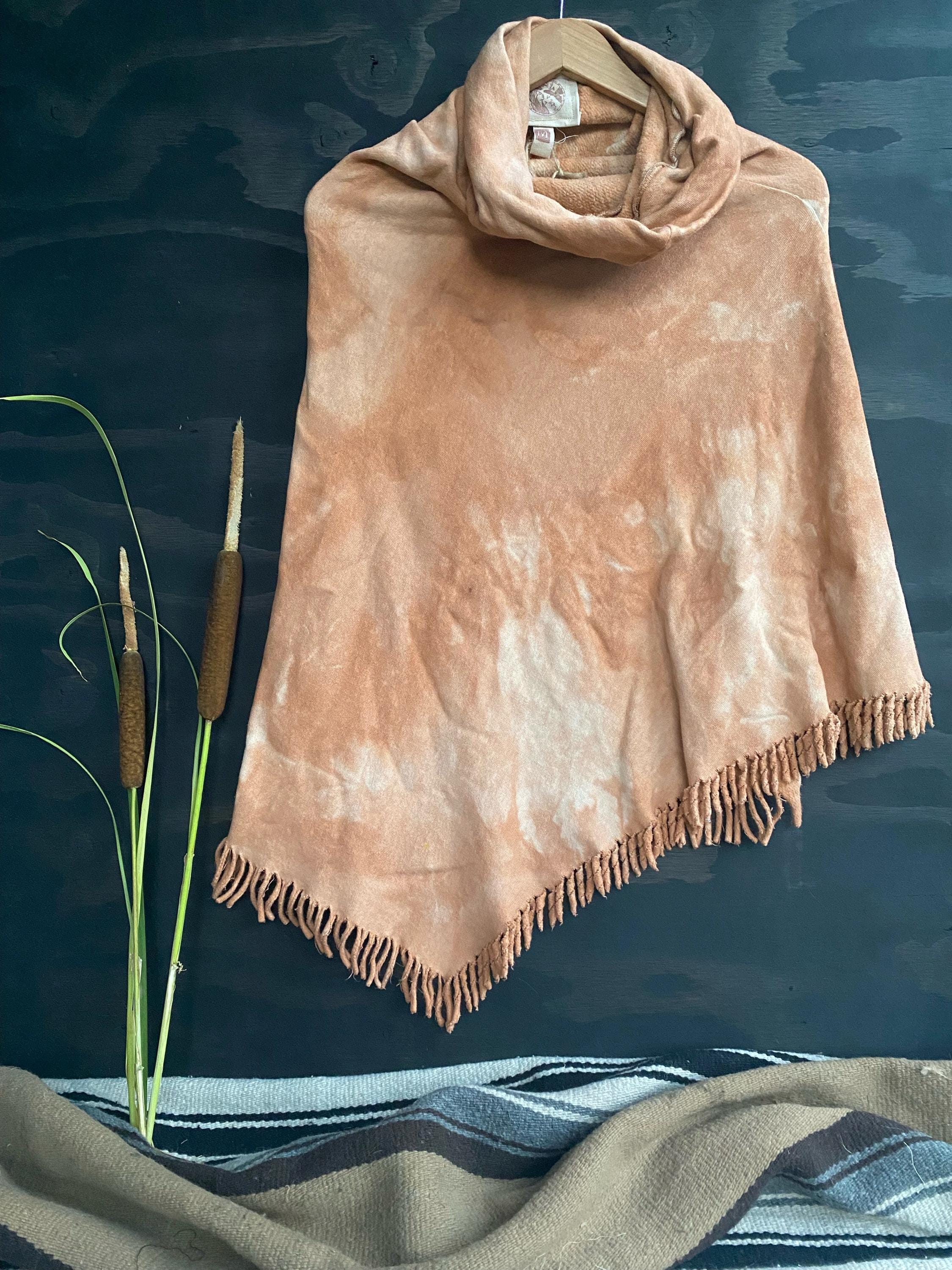 A terracotta-colored hemp fleece poncho with a turtle neck and fringe detailing, displayed on a dark background with natural dyeing methods suggested by the text.