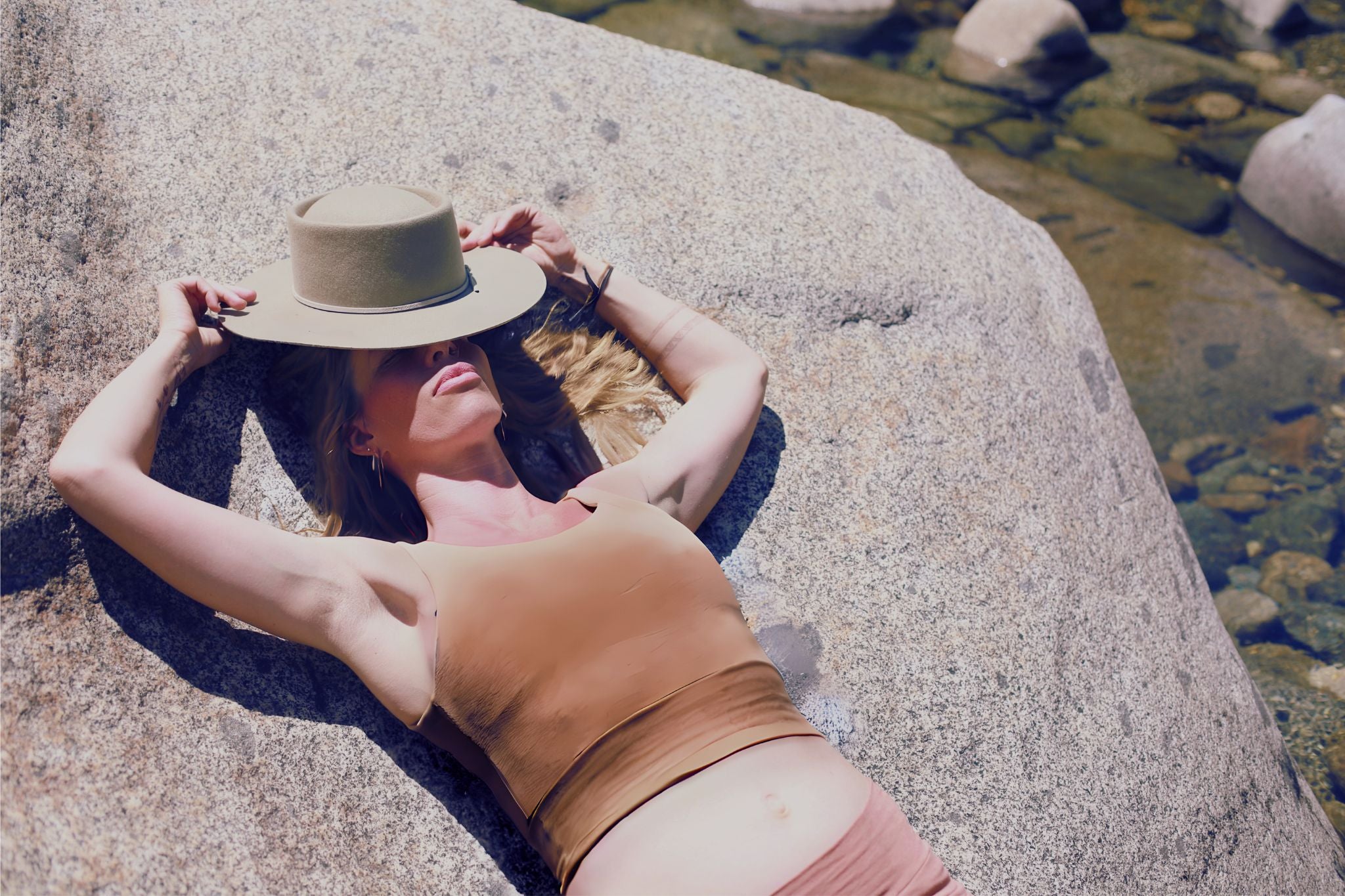Woman lying on a rock by a river bank wearing a hat and a top.